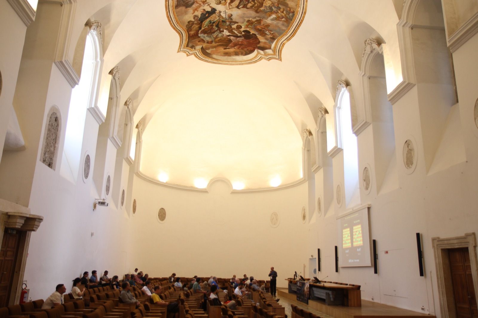 A session of AES 2026 in the main hall: rows of seated participants under a frescoed vault, the speaker at the lectern