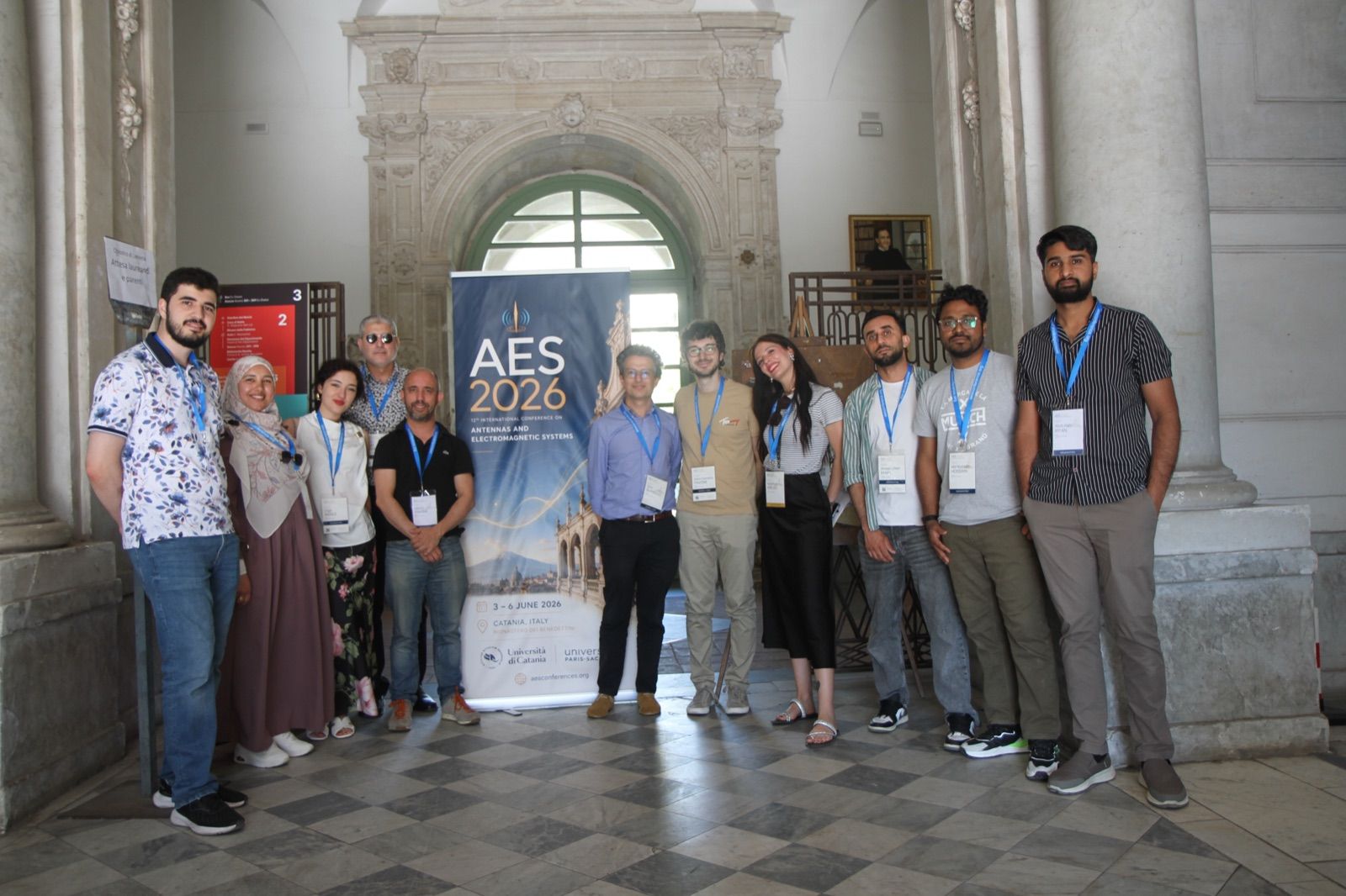 A group of AES 2026 participants standing together in front of the conference banner in the entrance hall