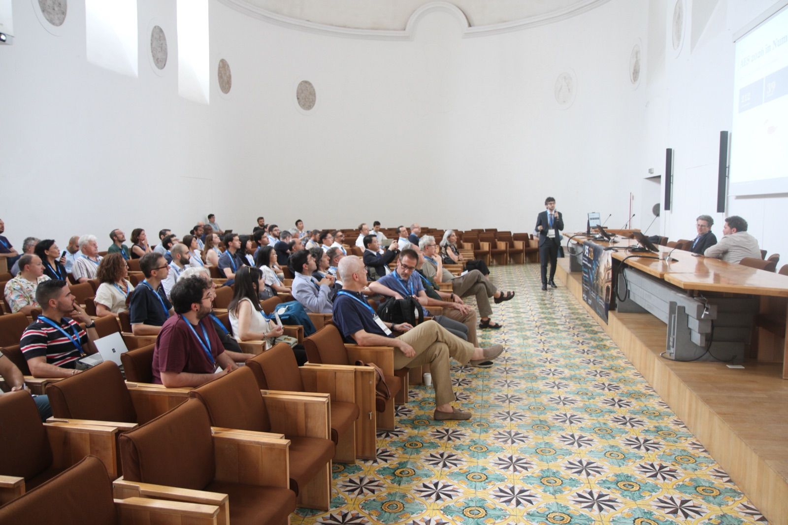 A conference session at AES 2026: the audience seated in a vaulted hall, a speaker at the lectern