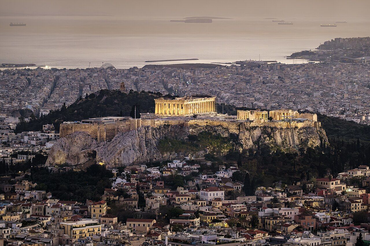 The Acropolis of Athens at dusk, its temples lit above the rooftops of the city, with the sea in the distance