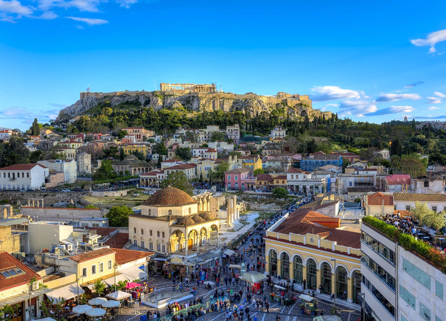 A square in central Athens seen from above, with its streets, rooftops and crowds, and the Acropolis on its hill beyond